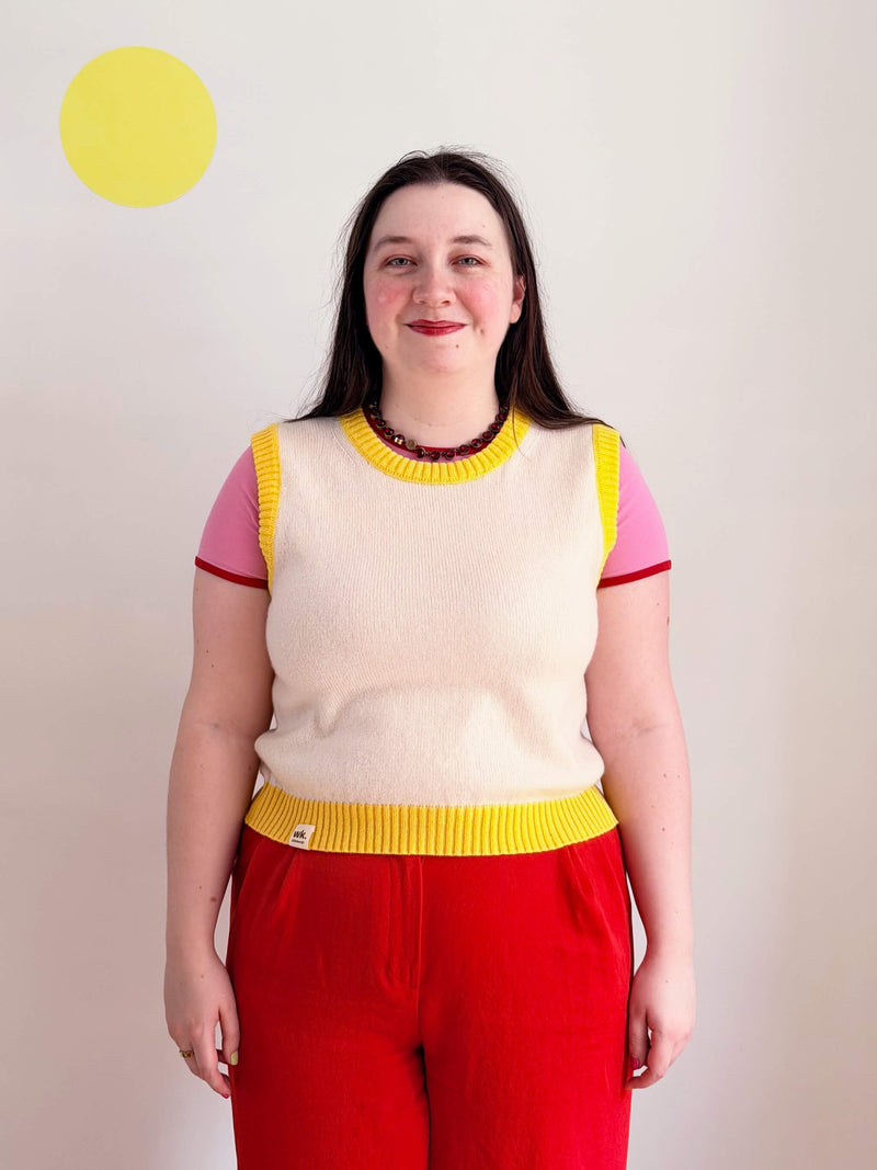 Person wearing a colourful yellow and cream sweater vest and red pants against a plain background