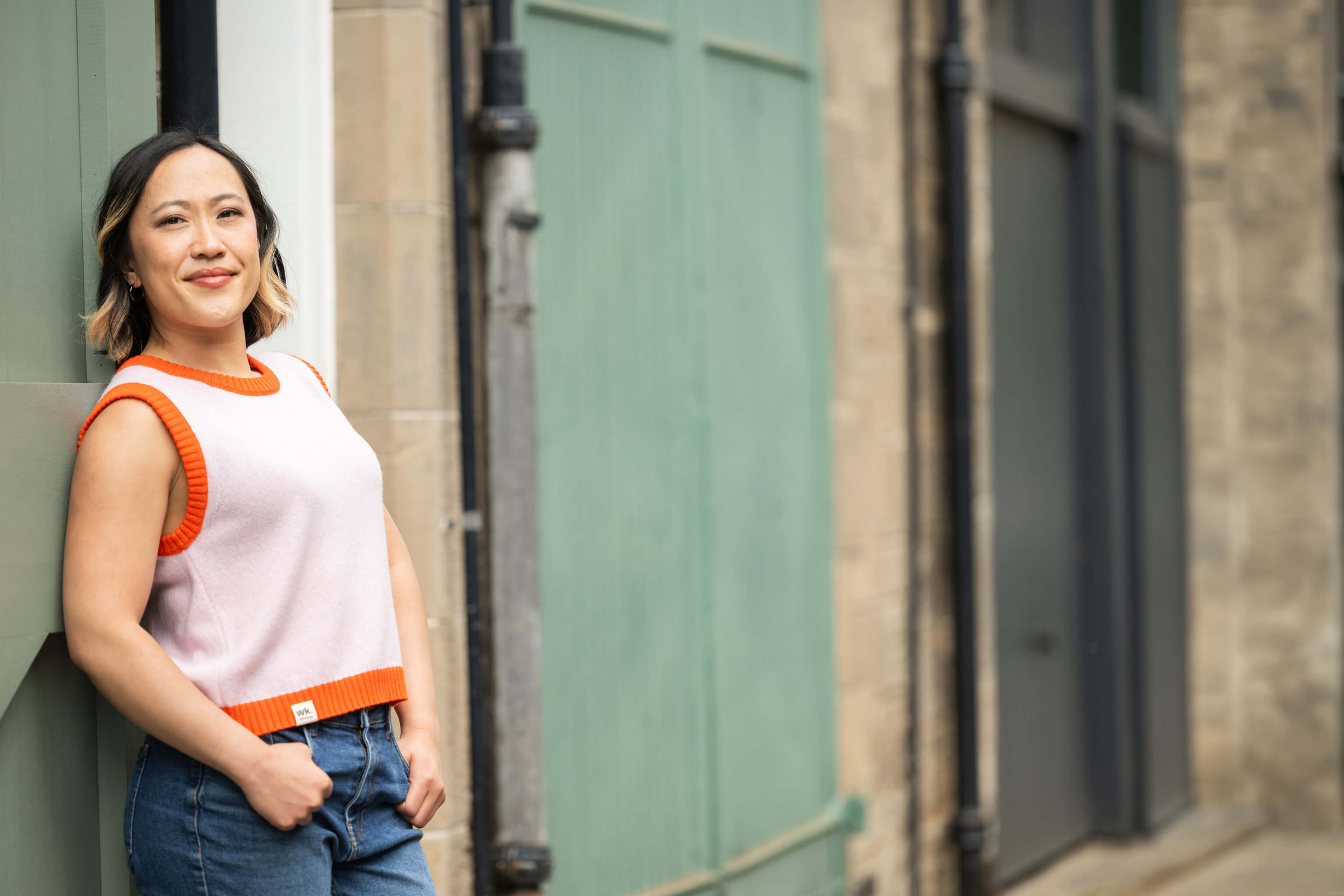 Woman in a white and orange sleeveless top leaning against a building.